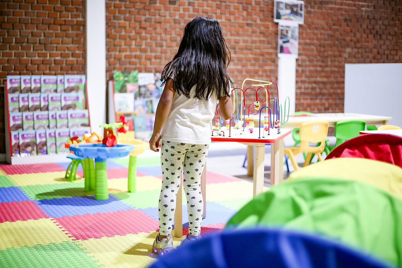 Niña de pie juega juguete de destreza, colocado encima de una mesa, al fondo se observan  un estante con libros y más mesas