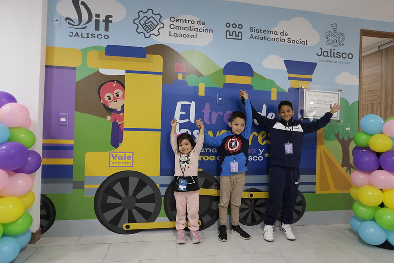 3 niños levantan las manos en la entrada de la ludoteca donde se observa el mural del tren de los valores